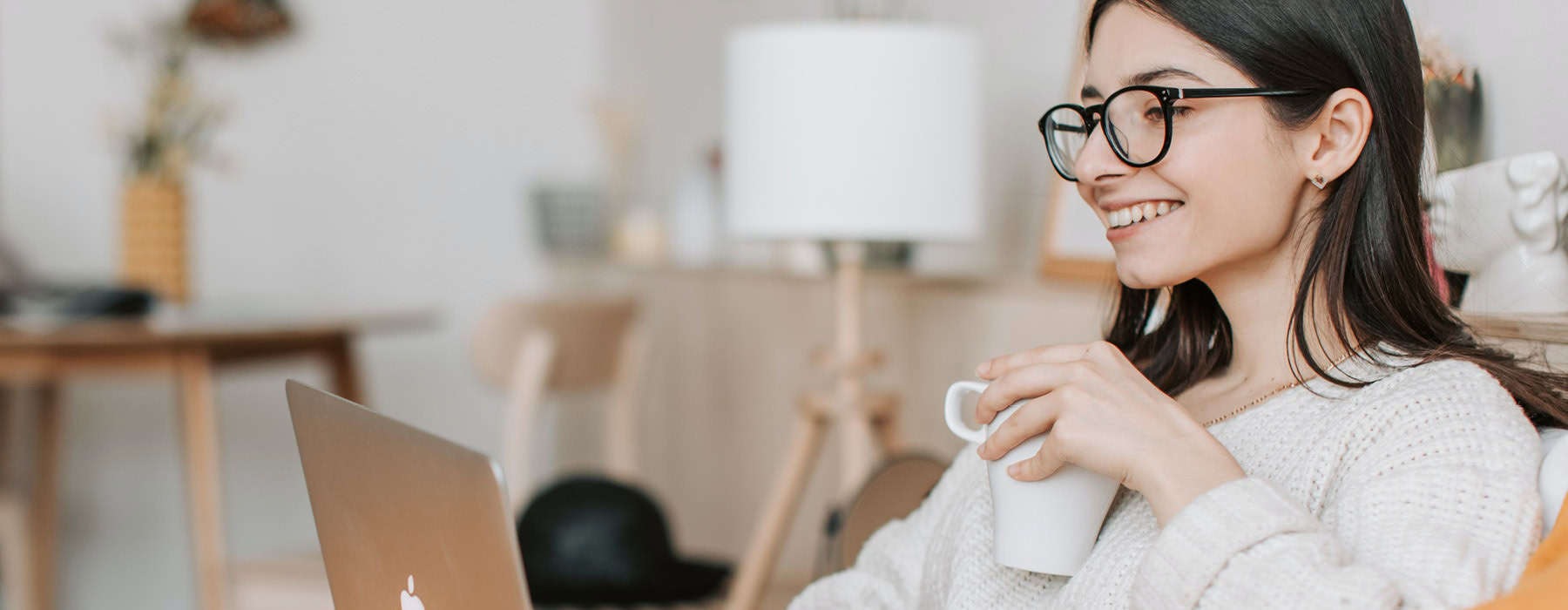 woman, with a cup of coffee, sits on her couch and looks at her laptop